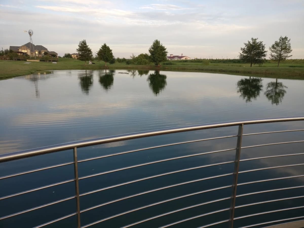 A pond with a fountain along a white ranch fence at Windsong Ranch in Prosper, TX.